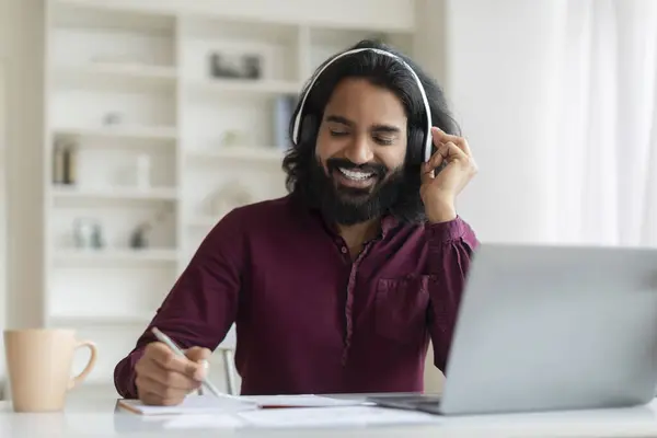 Cheerful young indian man working from home office, sitting at desk, using laptop and headset, smiling eastern male taking notes during video conference with colleagues, copy space