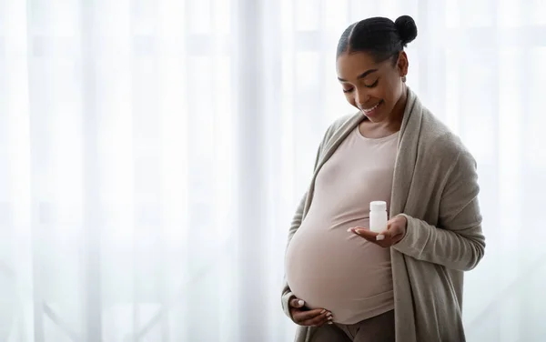 将来の母親のための彼女のサプリメントを示す美しい妊娠中の黒人女性は 彼女の大きなおなかと笑顔を抱き 家の窓の横に立って スペースをコピーします 妊娠中の栄養 健康的なライフスタイル — ストック写真