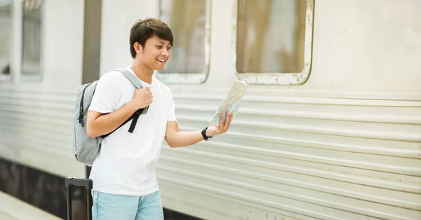 Young asian guy carrying backpack tourist checking paper map, standing by train at station, looking at destination point, panorama with copy space. Traveling, tourism, vacation