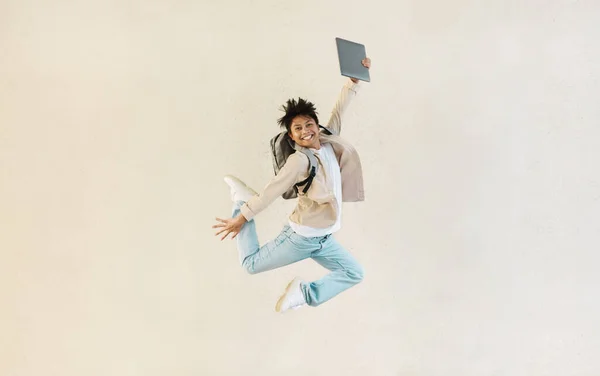 Remote job opportunities, freelance. Carefree millennial chinese man with computer laptop in his hand jumping up, carrying backpack, isolated on beige studio background, copy space