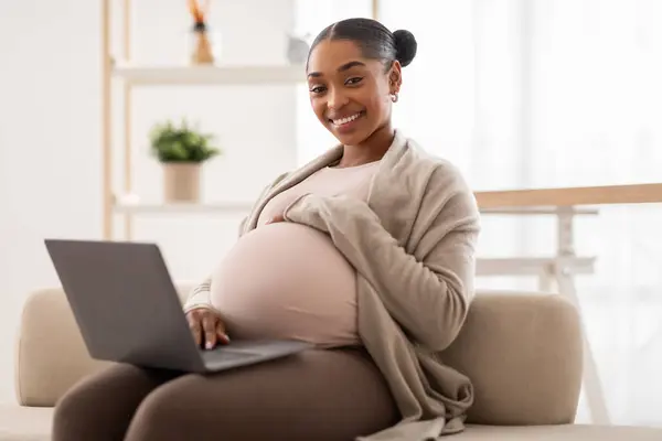 迷人的怀孕的非洲女人用笔记本电脑放松身心 上网浏览或与朋友聊天 阅读怀孕博客 坐在家里沙发上 空荡荡的空间 — 图库照片
