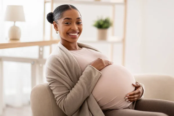 美しい妊娠中の黒人女性が彼女の腫瘍を抱きしめ 彼女の妊娠 自由なスペースを楽しんでいます アフリカ系アメリカ人のサイドビュー 自宅でソファに座って彼女の大きな腹に触れる女性を期待 — ストック写真