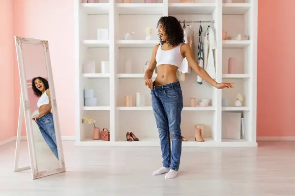 Exuberant curly-haired woman celebrating weight loss by fitting into oversized jeans, in a bright room with a full-length mirror and pastel decor