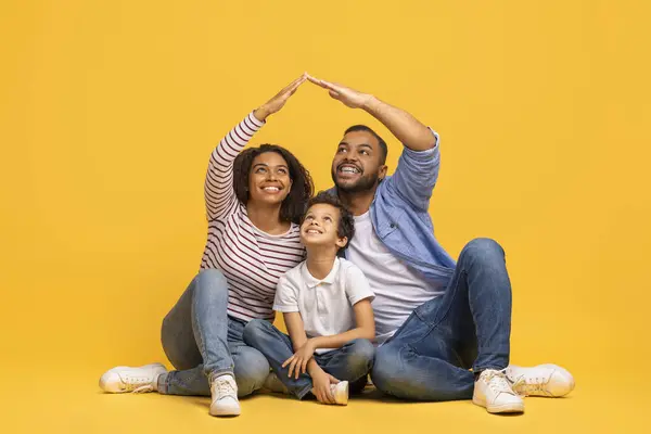 美しい黒人家族が床に座って 家の象徴 陽気なアフリカ系アメリカ人の両親と 黄色の背景にポーズする小さな息子 コピースペースを作ります — ストック写真