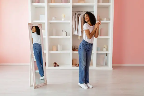 Joyful woman in casual jeans and white t-shirt smiling at her reflection in a full-length mirror in a bright, minimalist dressing room, full length