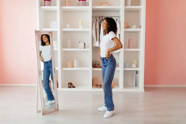 Radiant woman in white top and blue jeans admiring her outfit in a full-length mirror in a room with a chic, pastel-colored interior