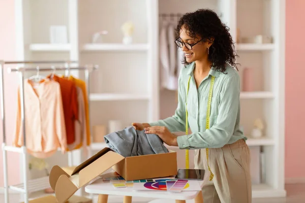 Joyful fashion designer with curly hair and glasses unpacking a box of new clothing designs in her sunny, pastel-colored design studio