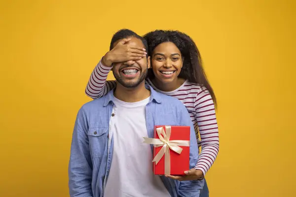 Surprise Gift. Romantic black woman covering her boyfriends eyes and giving present, caring lady greeting husband with birthday, wedding anniversary or valentines day while standing on yellow