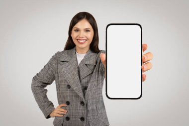 Gri kareli ceketli neşeli iş kadını cep telefonunu boş ekranla sunuyor. Uygulamalar ve reklamlar için mükemmel.