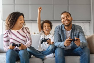 Aile eğlencesi. Neşeli siyahi ebeveynler evde oğluyla video oyunu oynuyorlar mutlu Afrikalı anne, baba ve erkek çocuk joystick kullanıyor ve birlikte eğleniyorlar.