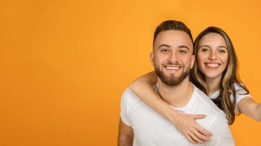 Kadını sırtında tutan mutlu, genç Avrupalı adam, eğleniyor, boş zamanlarının tadını çıkarıyor, turuncu arka plan stüdyosunda izole halde. Olumlu yaşam tarzı, aşk, romantizm ve ilişki, özçekim