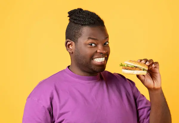 微笑的黑人手里拿着巨大的美味汉堡 微笑的相机站在黄色的工作室背景上 与汉堡包 广告垃圾食品搭配的垃圾食品食客的肖像 — 图库照片