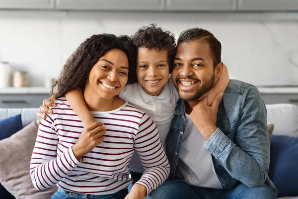 Happy Family Portrait. Smiling black parents and their little son posing together, embracing and looking at camera, african american man, woman and child bonding at home, sitting on couch
