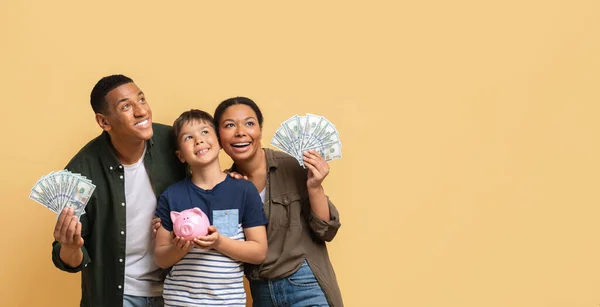 Family savings, budget planning, childrens pocket money. Positive loving young black father mother and preteen kid son holding cash dollar banknotes and piggy bank, looking at copy space, banner
