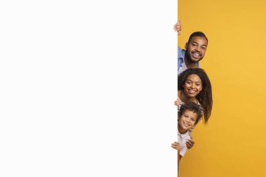 Beyaz reklam panosunun arkasından bakan üç kişilik neşeli siyah aile, mutlu bir ebeveyn ve genç oğul sarı arka plana karşı duruyorlar, yeni bir teklif öneriyorlar, kopyalama alanı.
