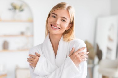 Beyaz bornozlu çekici sarışın bayan kendine sarılıyor, şımartma ve kendini kabullenme anından zevk alıyor, modern banyo düzeninde güzellik ve refahı yansıtıyor.