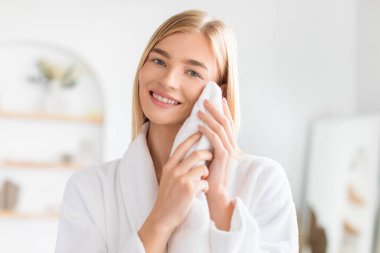 İçerde yıkandıktan sonra temiz yüzünü yumuşak havluyla kurulayan, modern banyoda bornozla duran, yüz hijyeninin ve cilt bakımının keyfini çıkaran, gülümseyen genç bayan.