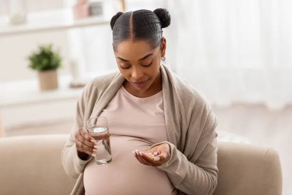 健康的年轻怀孕的非洲裔美国妇女 手里拿着药丸 一杯水 期待着女士在怀孕期间服用日常药物维生素补充剂 坐在家里沙发上 抄袭空间 — 图库照片