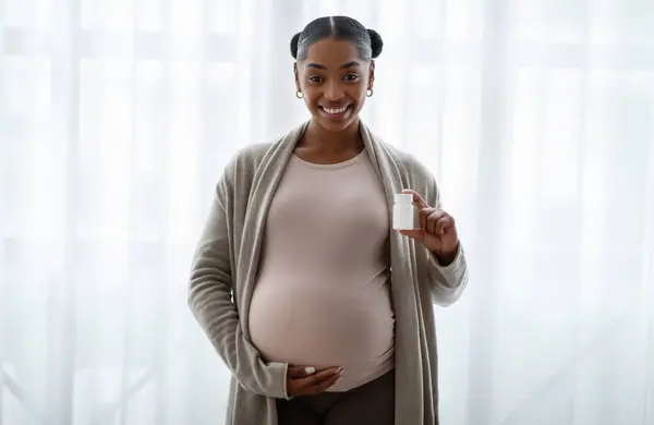 ポジティブなアフリカ系アメリカ人の妊婦はビタミンと白いプラスチック瓶を示し 母親を期待するためのサプリメント 自宅の窓の隣に立って 彼女の大きな腹を抱擁 — ストック写真