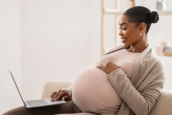 妊娠中のアフリカ系アメリカ人女性 コンピュータラップトップで自宅でリラックスし リビングルームでソファーに座り 妊娠ブログを読み オンラインで赤ちゃんのためのものを購入 サイドビュー コピースペース — ストック写真