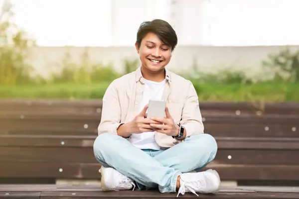 Smiling handsome young chinese guy wearing casual clothing using smartphone, chatting with friends, scrolling on social media, chilling at park, sitting on bench, copy space