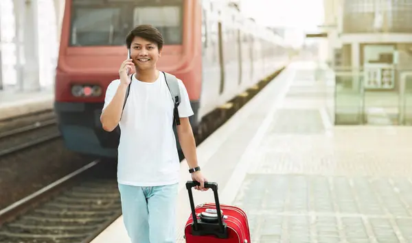 Cheerful young asian guy wearing comfy casual outfit walking by train station, carrying suitcase, talking on phone, calling taxi, booking hotel, copy space for advertisement, panorama