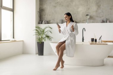 Beyaz bornozlu gülümseyen genç Hintli kadın duştan sonra akıllı telefon kullanıyor, banyoda küvette oturuyor, dişlerini fırçalıyor, telefonu tutuyor, sosyal ağları kontrol ediyor, mesajları okuyor, iyi haber.