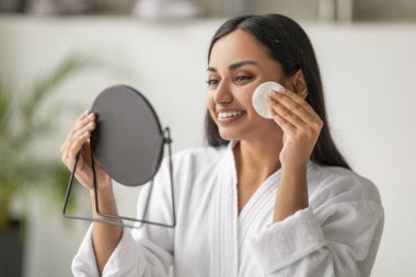 Neşeli, uzun saçlı, beyaz bornoz giyen, aynaya bakıp pamuk ped kullanan, sulandırıcı toner, deniz suyu, losyon ya da asit pedleriyle cilt temizleyen bir kadın.