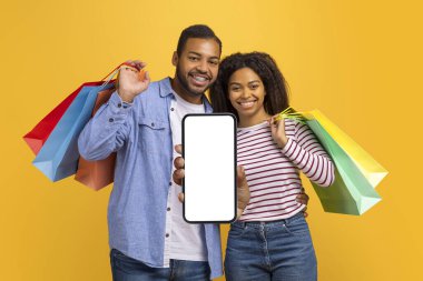 İnternetten alışveriş uygulaması. Neşeli genç siyah çift kameraya boş beyaz ekranlı akıllı telefon gösteriyorlar ve sarı stüdyo arka planında izole bir şekilde dururken ellerinde parlak alışveriş torbaları tutuyorlar.