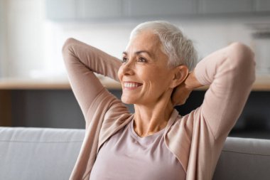 Gülümseyen olgun bir kadın kanepeye yaslanıp el ele tutuşuyor, hafta sonları sakin bir şekilde dinleniyor, evdeki modern oturma odasında kanepede oturuyor. Barışçıl mola kavramı