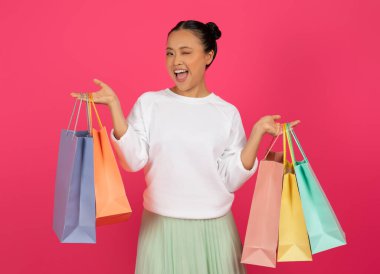 Neşeli Asyalı genç bayan elinde renkli alışveriş çantaları ve göz kırpan, mutlu Koreli alışveriş hastası bayan parlak pembe stüdyo arka planında eğleniyor, sezonluk satışlardan ve indirimlerden keyif alıyor, bedava alan bir yer.