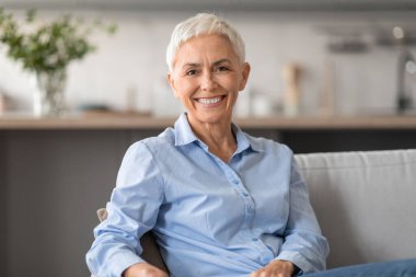 Kısa gri saçlı, neşeli, kendine güvenen beyaz kadın portresi, rahat bir koltuğa oturmuş kameraya gülümsüyor, modern salonda kendine güven ve pozitiflik yayıyor.