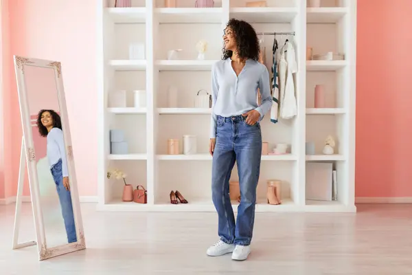 Smiling curly-haired woman in a chic blue blouse and casual jeans looks pleased with her reflection in a full-length mirror in a pink room