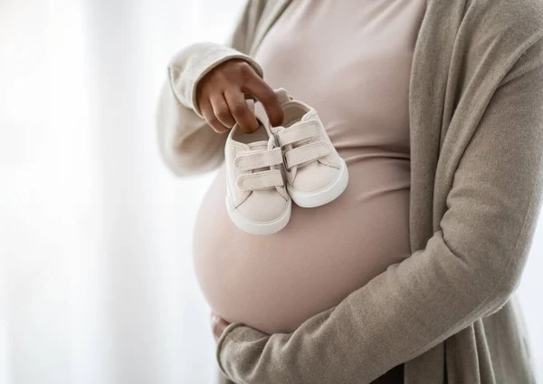 自宅の窓の隣に立っている妊婦の腹の上の胎児のための小さな靴のクローズアップ 子供は 期待する女性の腹の未生児のためのバラバラ コピースペース クロップ — ストック写真