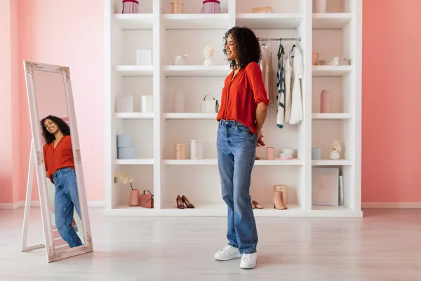 Smiling woman wearing red blouse and blue jeans, admiring her reflection in a full-length mirror in a room with pink walls and white shelves