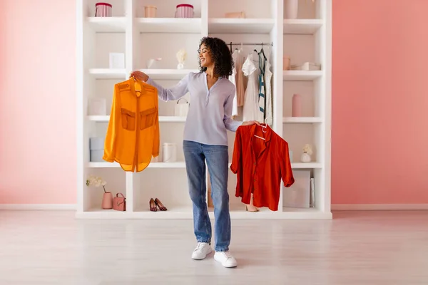 Happy curly-haired latin woman holding yellow and red shirts, comparing them for her outfit in a chic closet within a room with pastel pink walls