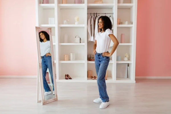 Confident curly-haired woman posing in a white t-shirt and jeans, looking at her reflection in a full-length mirror in a stylish room