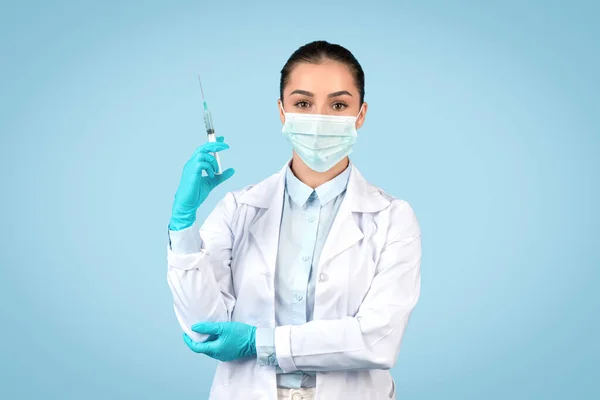 European lady doctor in white coat and surgical mask holding syringe, preparing for vaccination or medical procedure, isolated on blue studio background