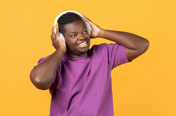 最喜欢的音乐 穿着便衣紫色T恤 头戴无线耳机的快乐的黑人男人 在线聆听着伟大的歌曲 在黄色工作室的背景下带着微笑看更远的地方 — 图库照片