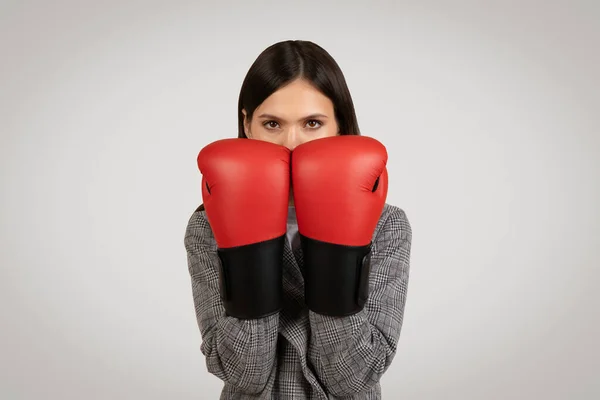 Resolute businesswoman with red boxing gloves covering her face, symbolizing challenge and competition in the corporate world, against grey background
