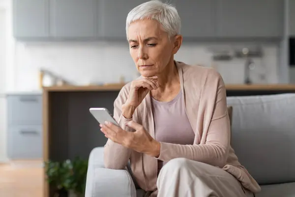 Doomscrolling. Serious Mature Woman Using Smartphone And Frowning Reading Bad News Online, Sitting On Couch At Home Interior. Technology And Communication Problems