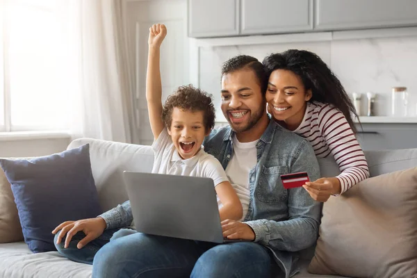 Happy Black Family With Laptop And Credit Card Relaxing At Home, Smiling African American Parents And Little Son Making Online Shopping While Resting On Couch In Living Room, Enjoying Sales