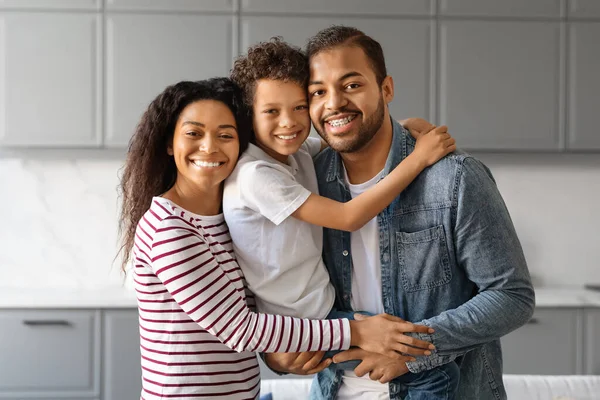 Happy black family with preteen son hugging and smiling in their kitchen, cheerful african american parents and male kid hugging together, posing in bright, modern home interior, free space