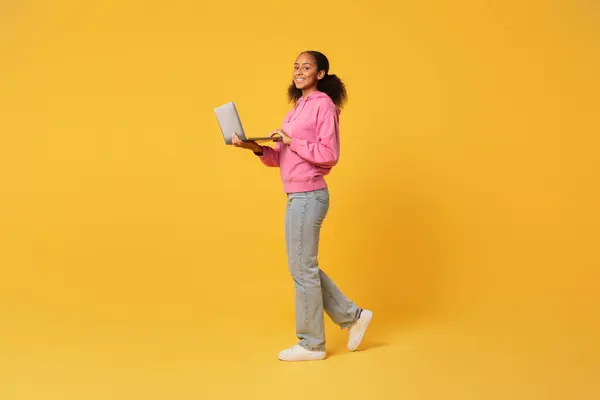 在工作室里 用笔记本电脑在黄色的背景下散步 微笑着的黑人女学生 全长拍摄的青少年在网上浏览 免费提供网上教育 — 图库照片
