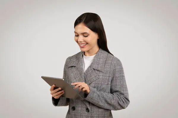 专注的年轻职业女性微笑着使用数码平板电脑 穿着别致的格子夹克 背景为柔和的灰色 — 图库照片