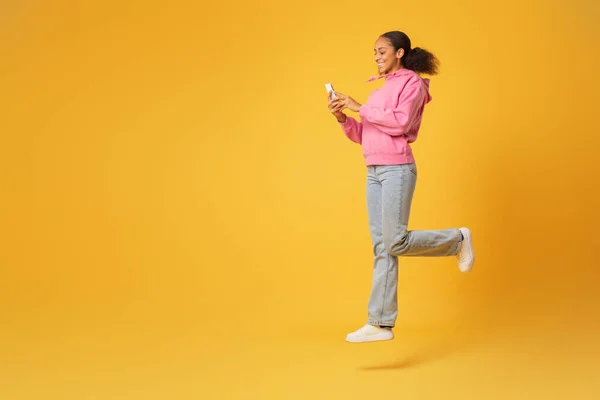 Happy African American student jumps in mid air browsing her smartphone, encapsulating youth and technology on yellow backdrop, surfing internet on gadget. Full length, free space