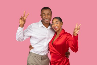 Heyecanlı Afrikalı Amerikalı çift zarif kıyafetler giyerek barış işareti yapıyorlar, pembe arka planda kaygısız titreşimler yayıyorlar ve kameraya gülümsüyorlar.