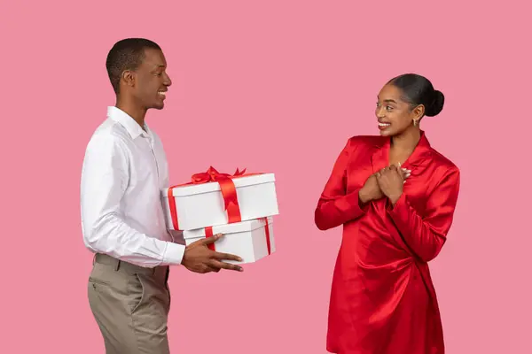 Generous black man in white shirt presenting large gift with bow to touched woman in red dress, her hands on her heart, against tender pink background