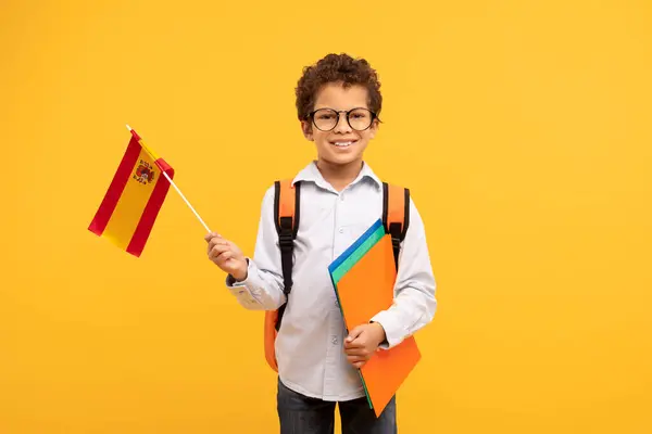 Proud student boy with curly hair and glasses holding Spanish flag and school folders, exuding confidence and readiness on yellow background