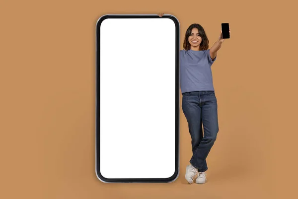 黒い空白のスクリーンが付いているスマートフォンを示す陽気な若いアラブの女性は 白い空のスクリーンのモックアップのコピー スペースが付いている大きい携帯電話の隣に オンライン提供 スタジオの背景を推薦します — ストック写真